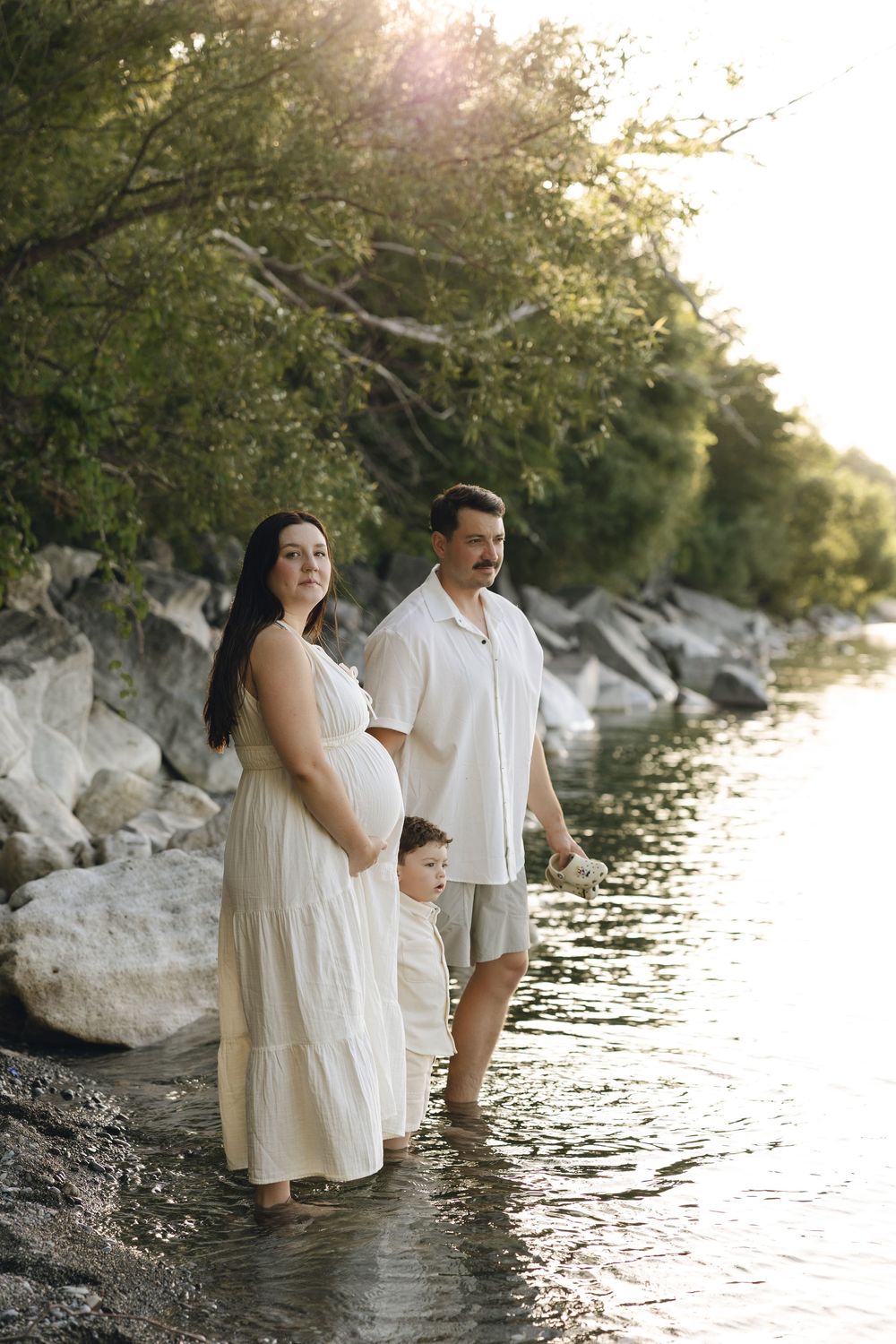 Three people in white clothing stand at waters edge during sunset, creating a serene family portrait against rocky shore.