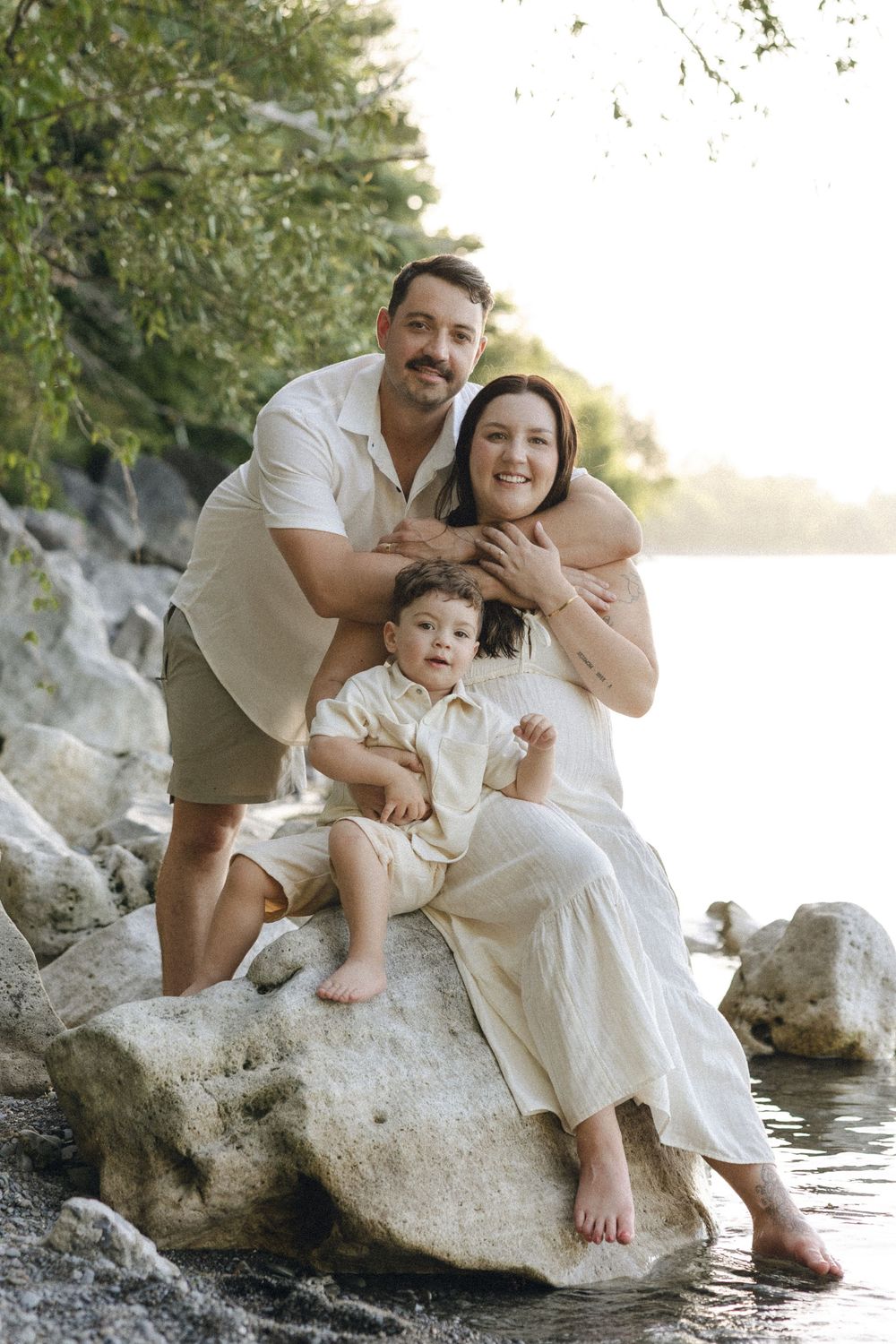 A family sits together on large rocks by the water's edge during a sunset photoshoot in neutral-colored outfits.