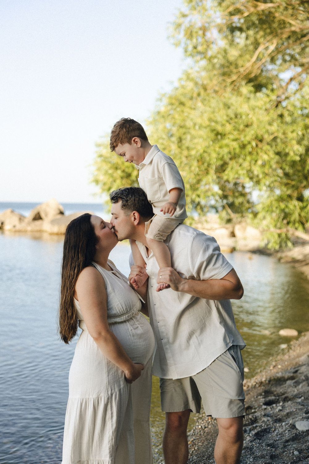 A couple shares a tender moment by a lake while their young child sits on shoulders during a sunset photoshoot.