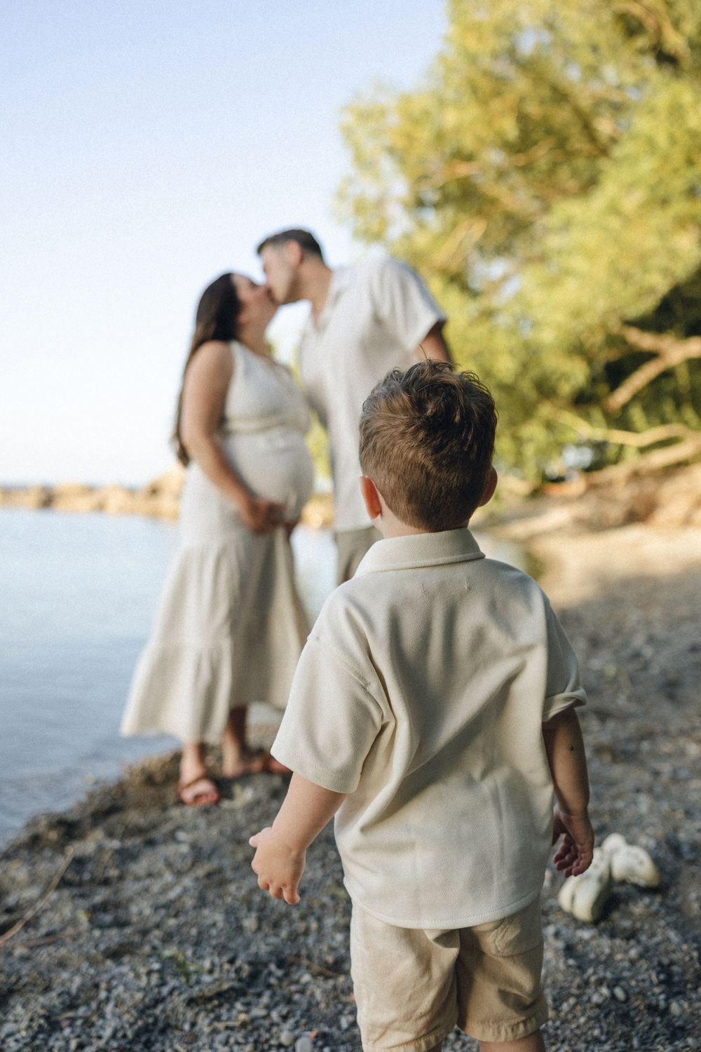 A toddler in beige clothing watches two figures distance on a rocky beach at sunset.