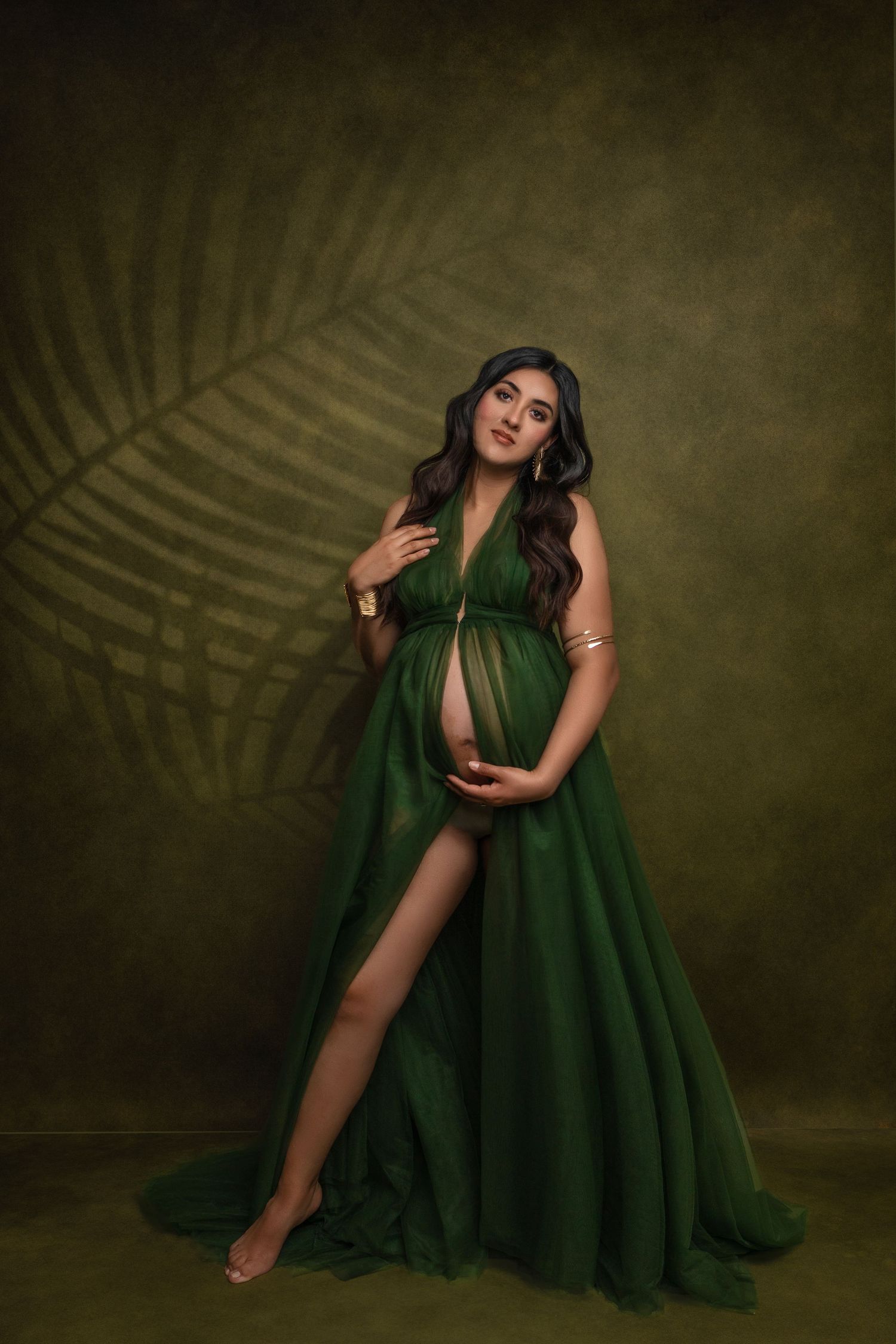 Artistic maternity photo featuring an elegant emerald green flowing gown against an olive backdrop with palm leaf shadow.