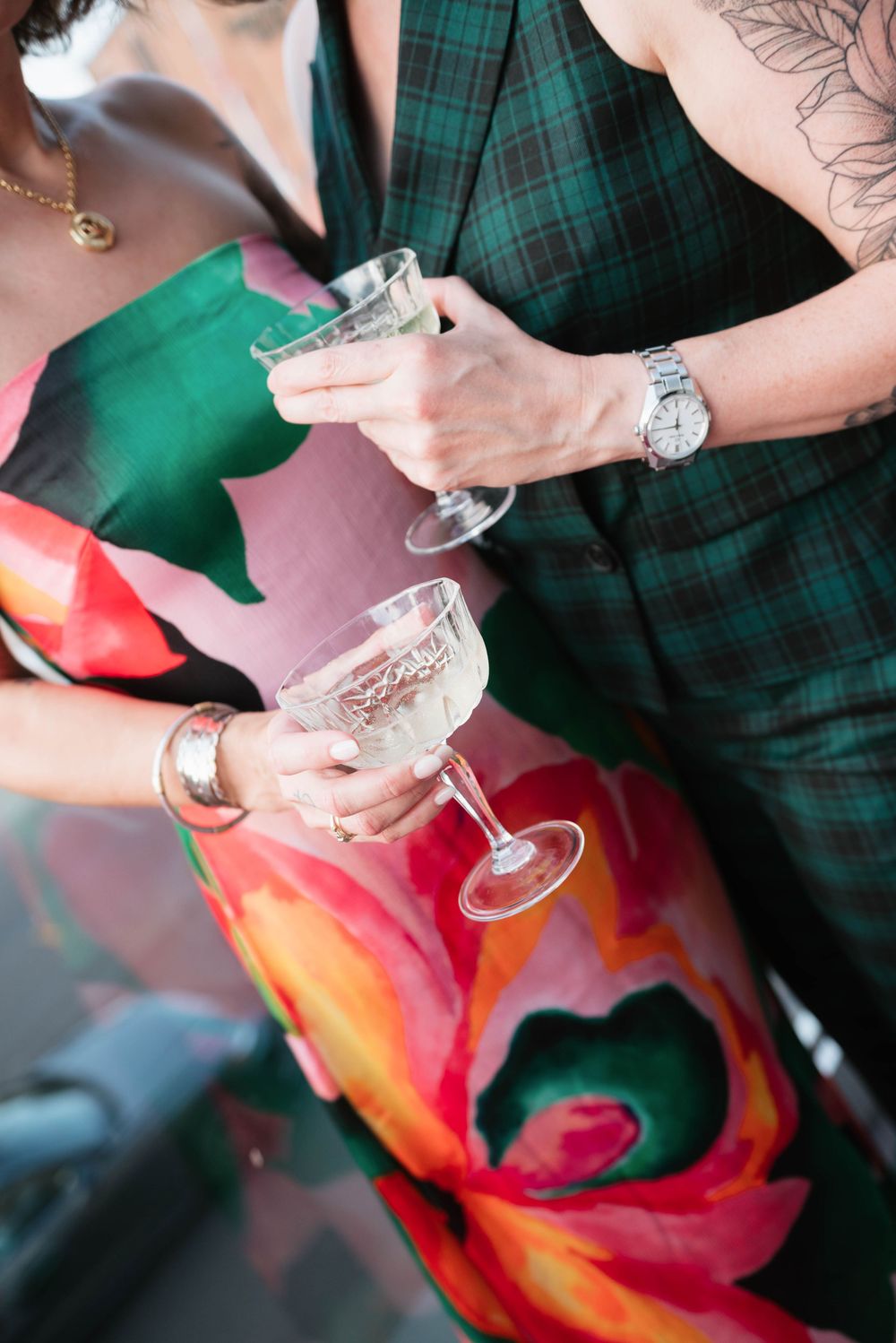 Two people clinking champagne coupes, wearing colorful floral and green plaid outfits, with jewelry and tattoos visible.