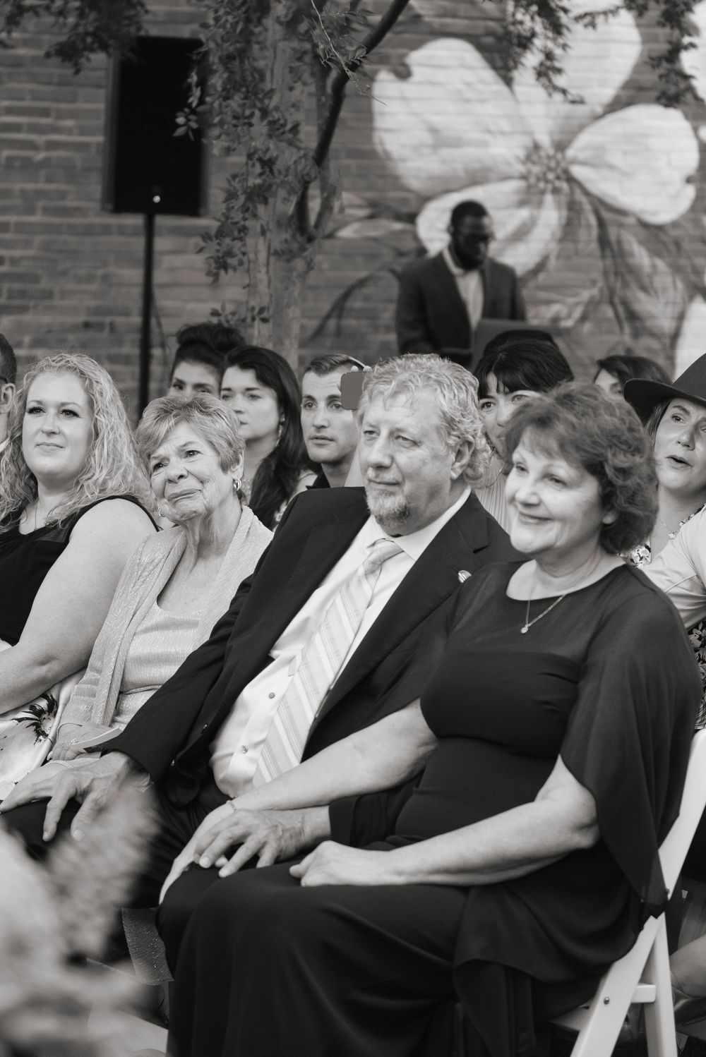 Black and white photo of wedding guests seated outdoors, smiling couple in foreground, floral mural on brick wall behind.