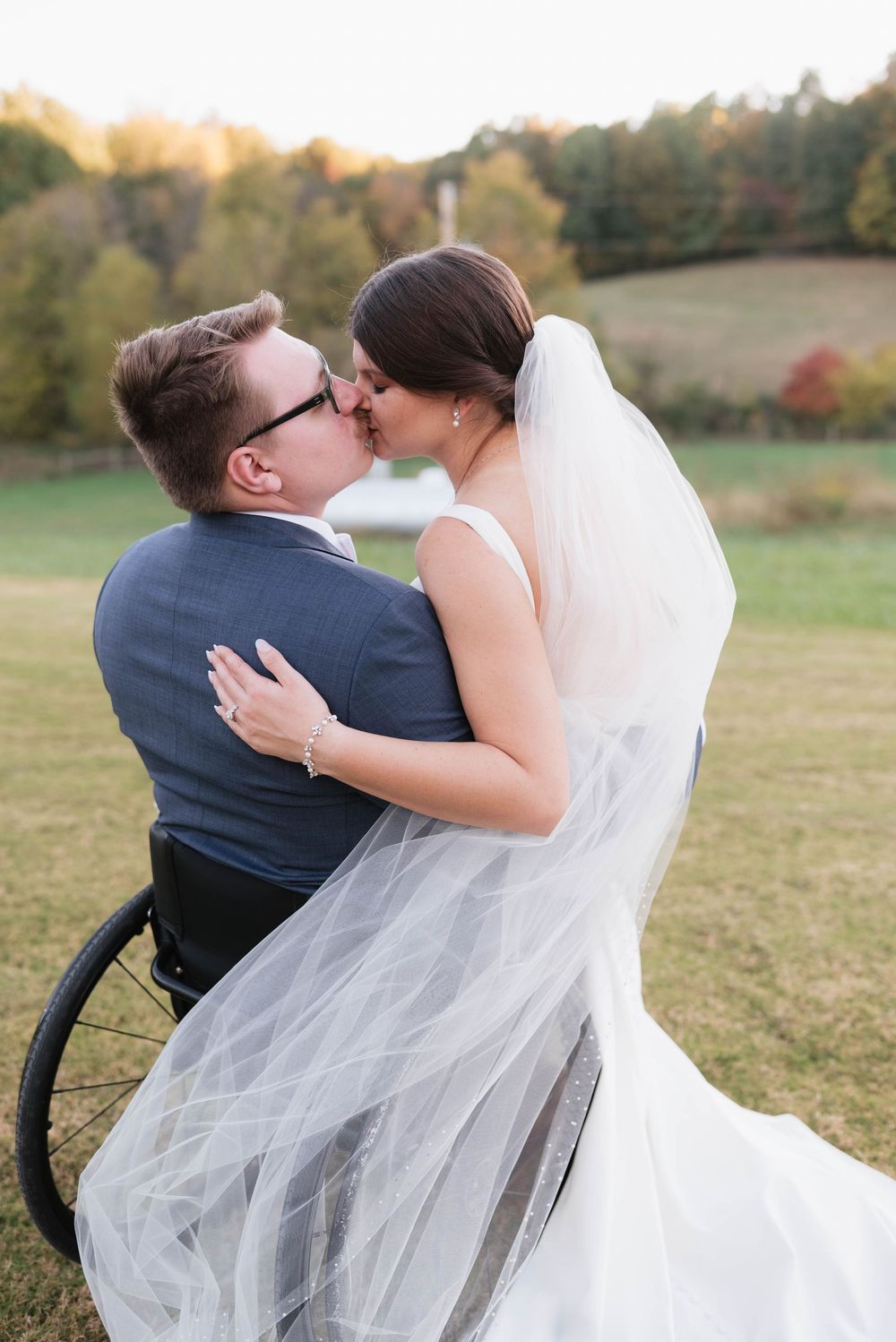 Bride in white gown kissing groom in wheelchair on grassy field during outdoor wedding ceremony.