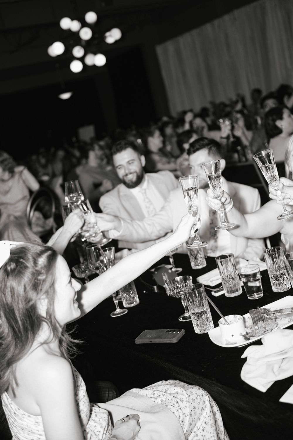 Black and white photo of guests raising glasses in a toast at an elegant wedding reception dinner.
