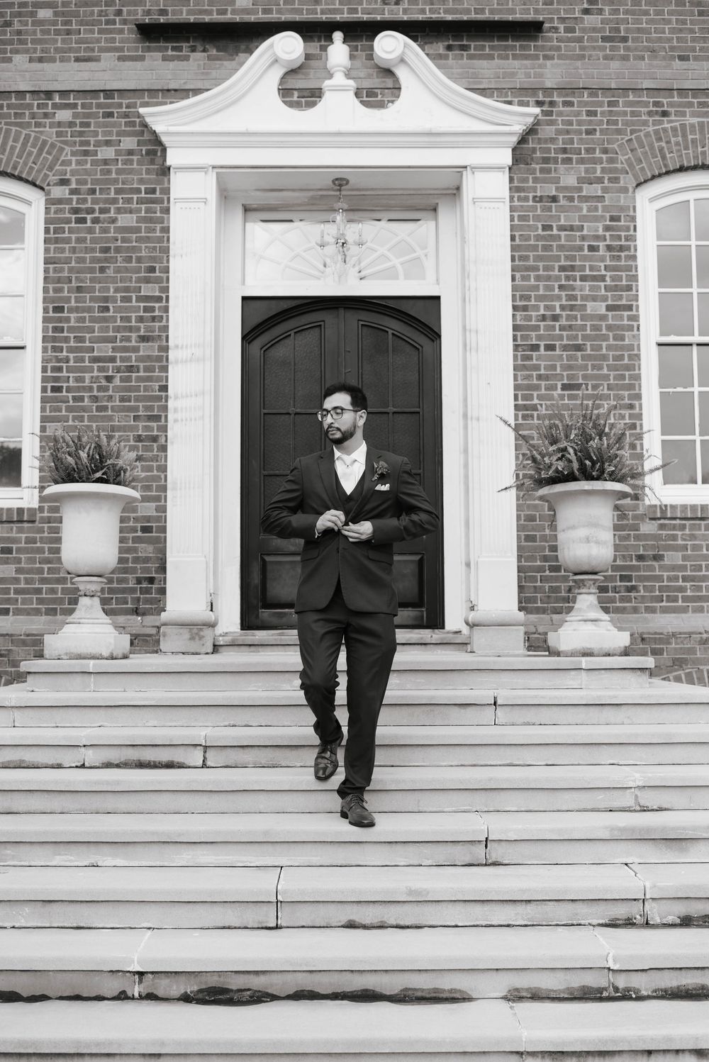 Groom in dark suit descending steps of elegant brick building with white columns and arched doorway.