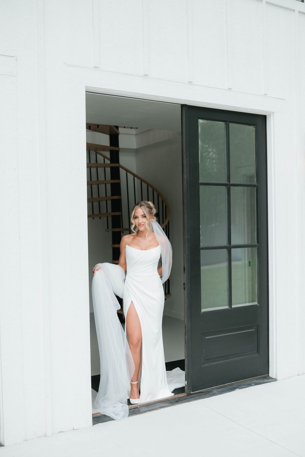 Elegant bride in sleek white gown with thigh slit posing in doorway of white barn venue.
