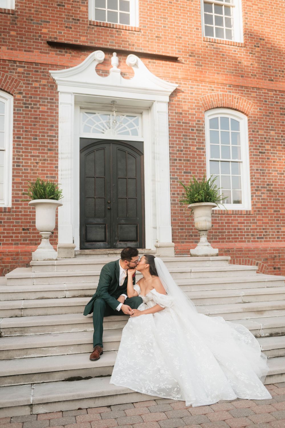 Bride and groom share a kiss on steps of elegant red brick venue, bride in lace gown with veil, groom in dark suit.