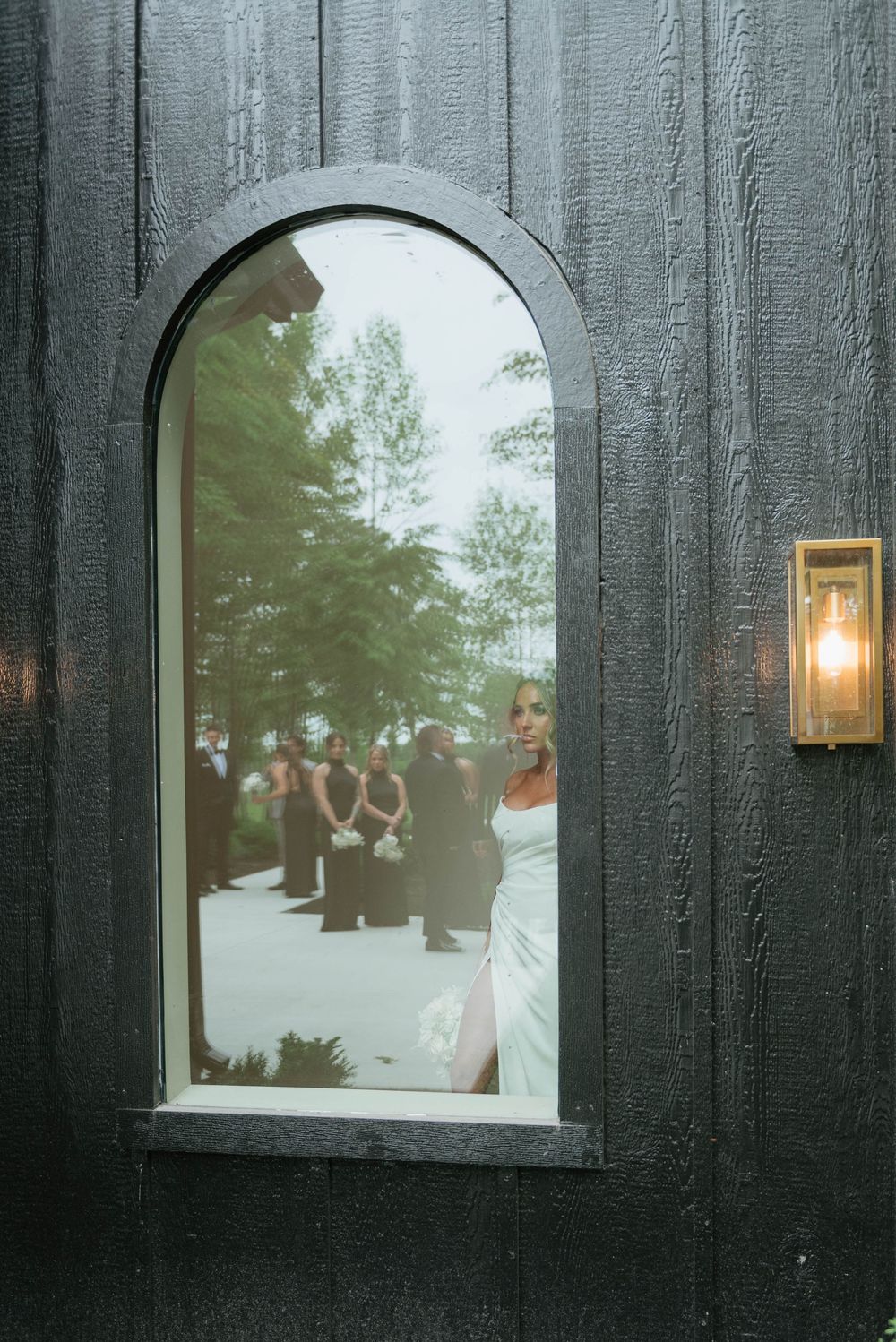 Bride in white gown viewed through arched window of dark wood door with guests in background.