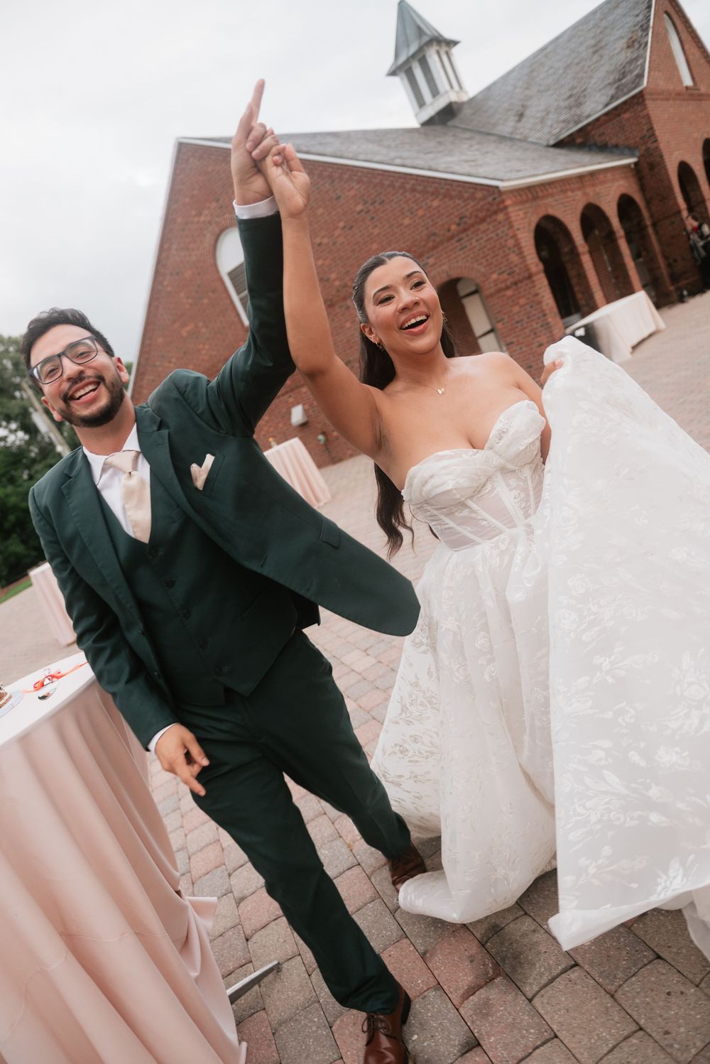 Joyful newlyweds celebrate outside a brick church, groom in green suit raises bride's hand as she laughs in a strapless lace gown.