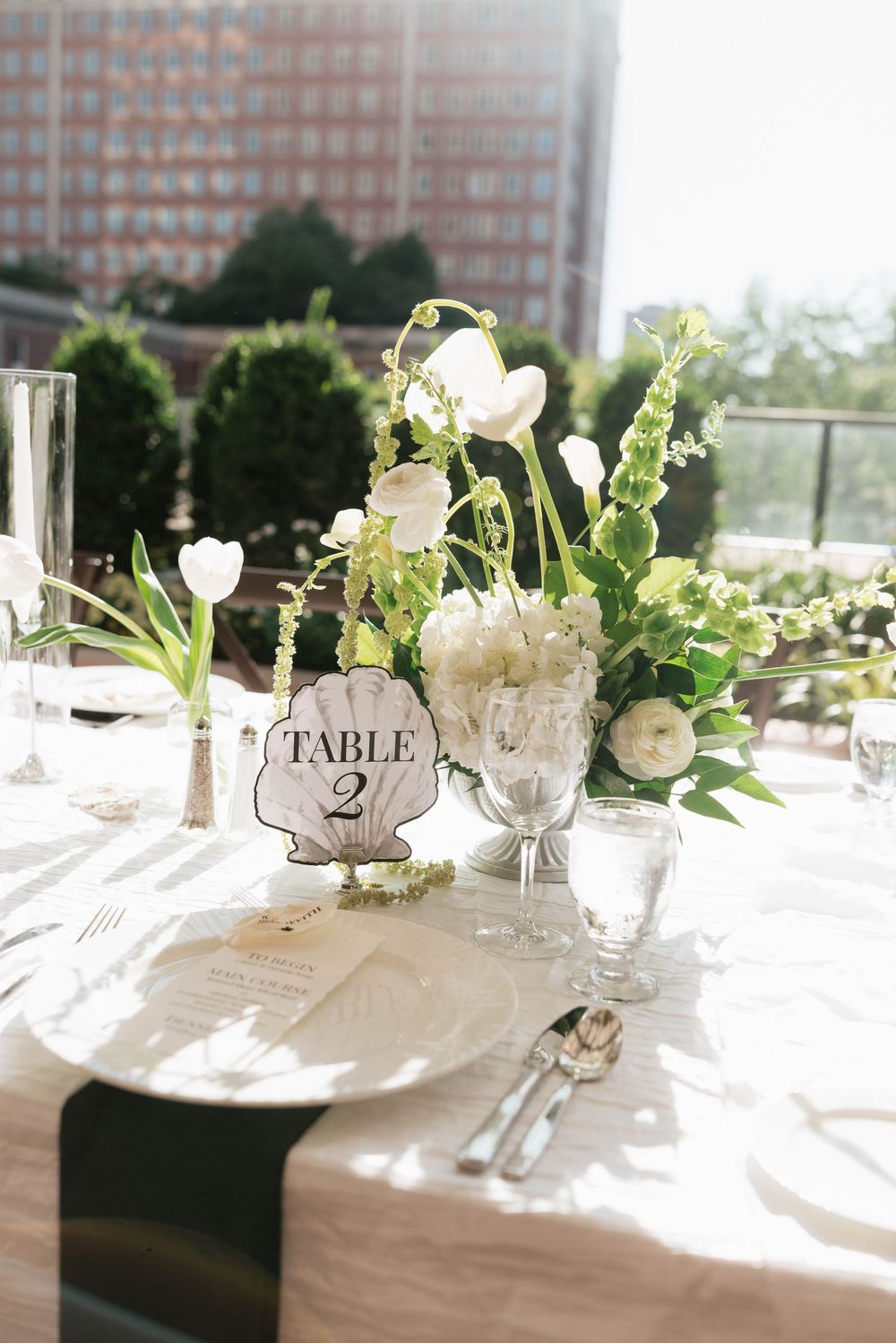 Elegant wedding reception Table 2 setting with white floral centerpiece, greenery, crystal glassware, and silverware on white linen.