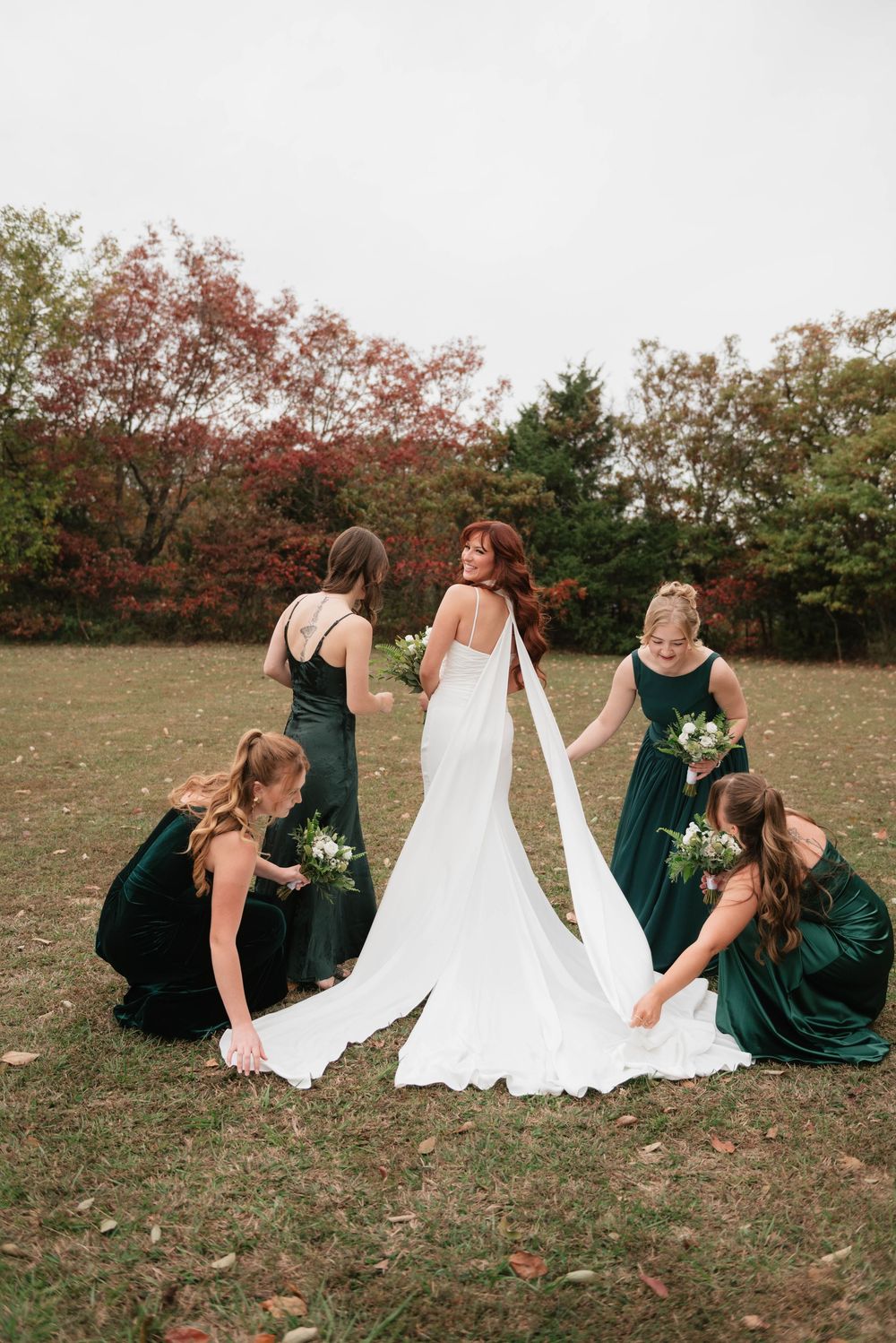Bride in white gown with bridesmaids in emerald green dresses arranging her train outdoors in autumn.