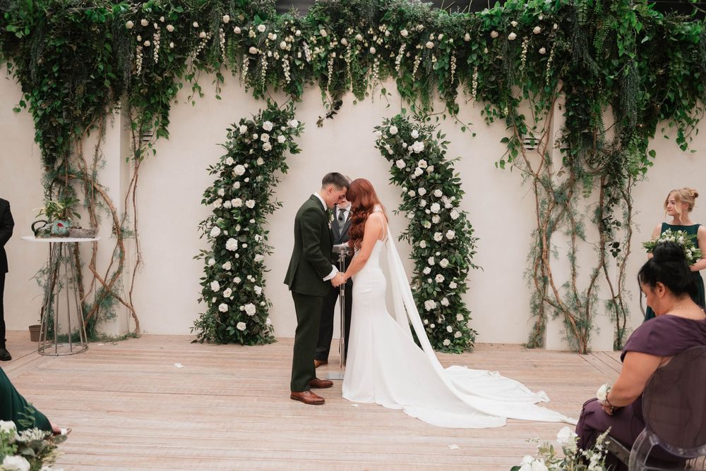 Couple exchanging vows under lush greenery and white floral arch during elegant indoor wedding ceremony.
