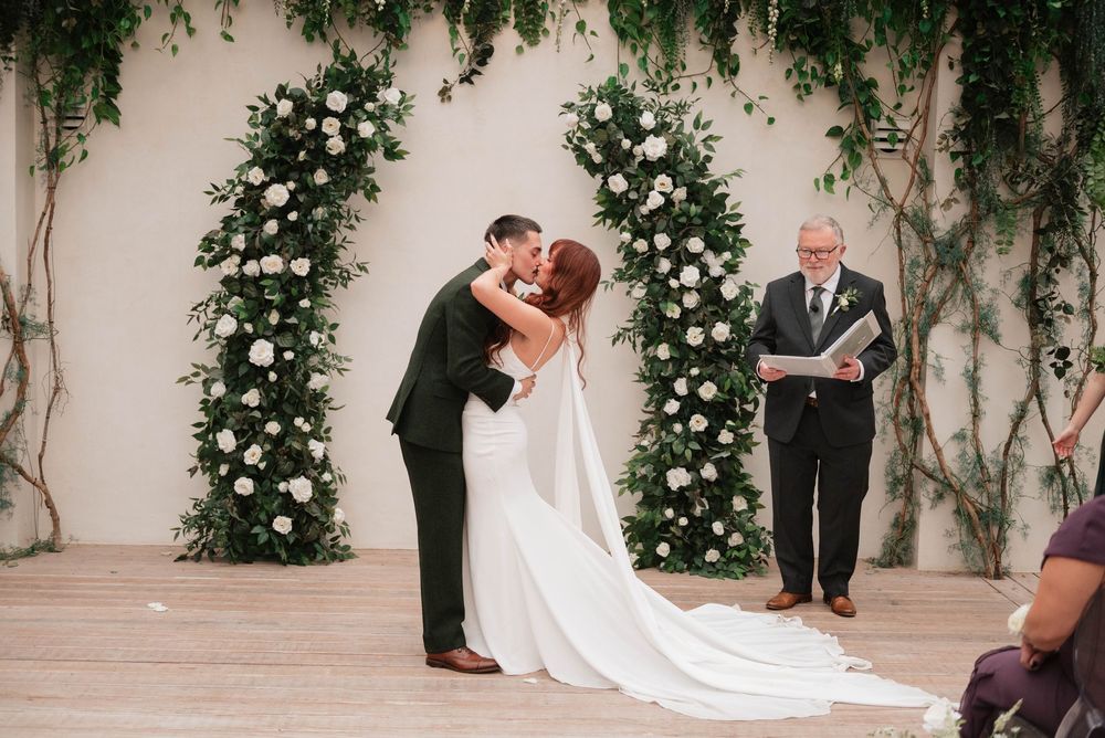 Bride and groom sharing their first kiss at the altar beneath a floral arch with white roses.