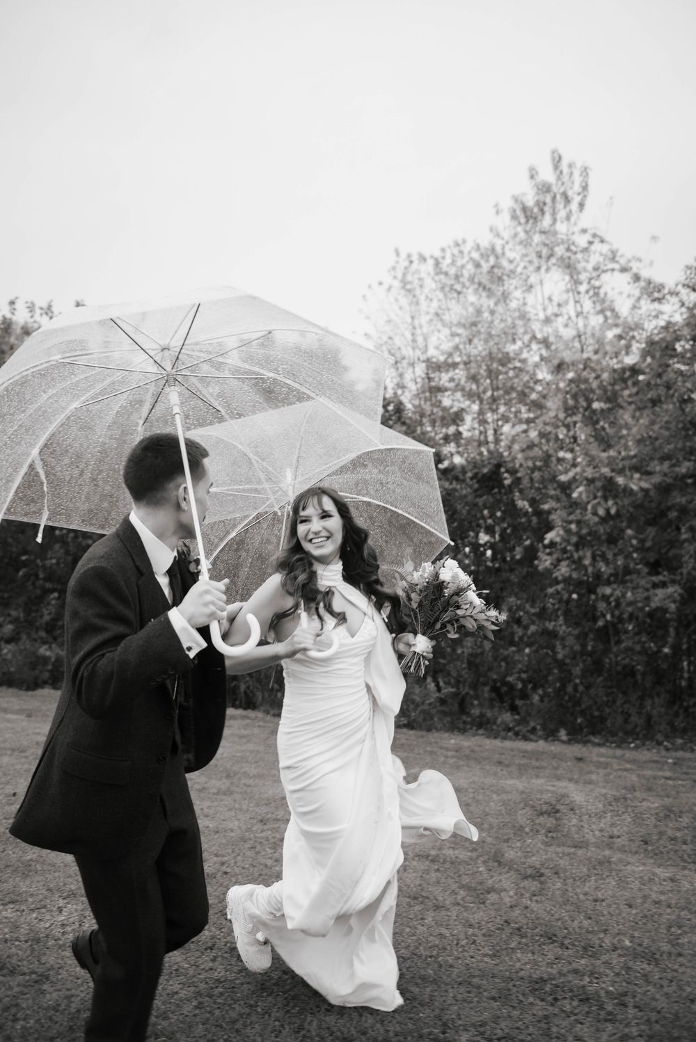 Black and white photo of joyful bride and groom walking with clear umbrellas on their wedding day.