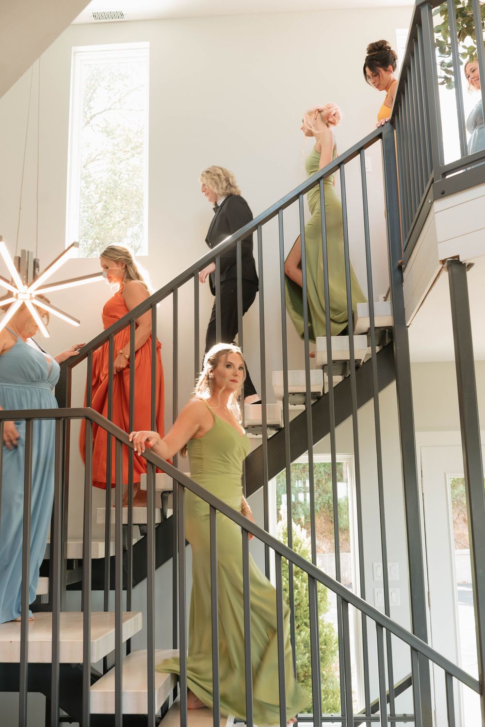 Bridesmaids in colorful gowns descend an elegant staircase during wedding preparations.