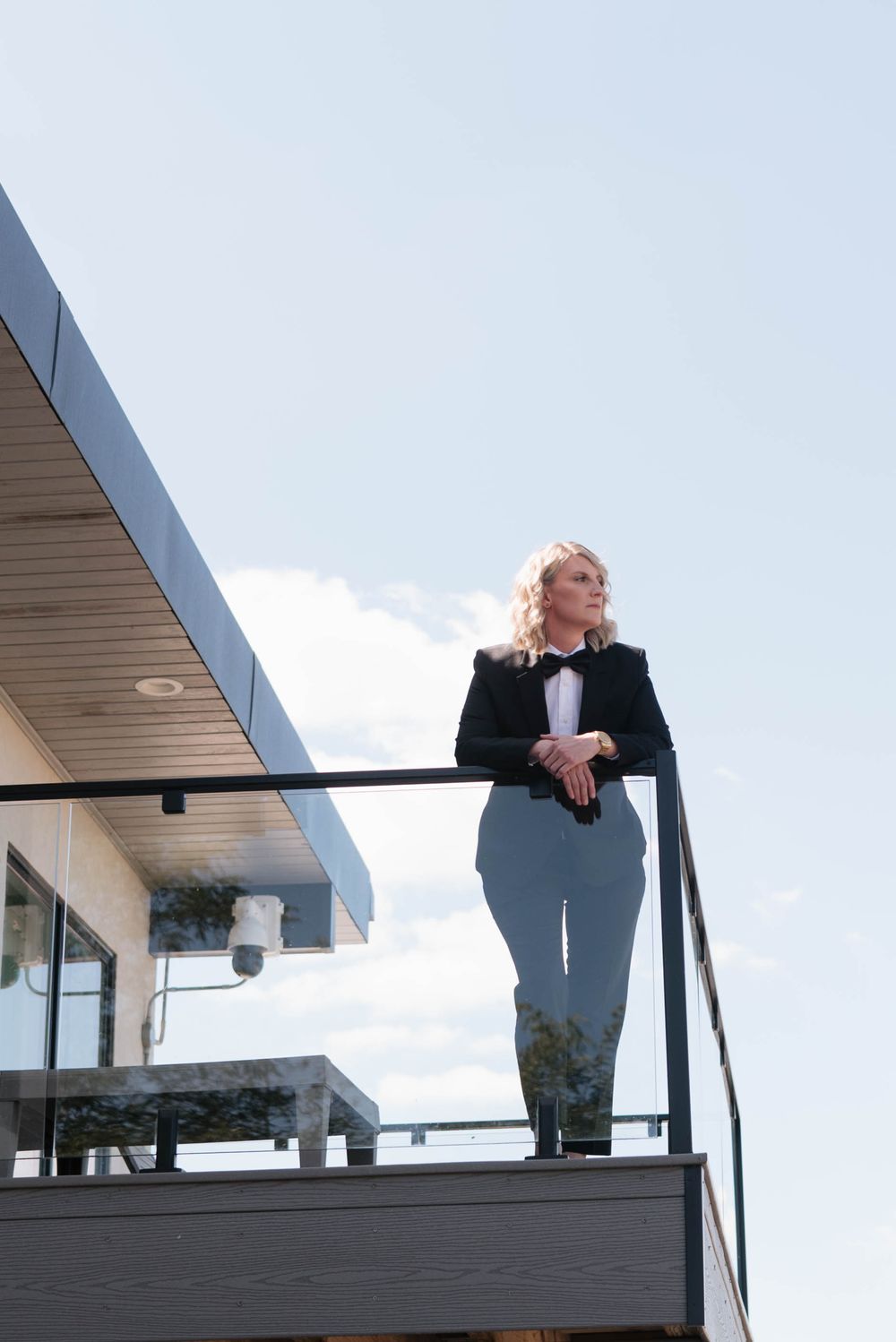 Elegant blonde woman in tuxedo standing on modern building balcony against blue sky.