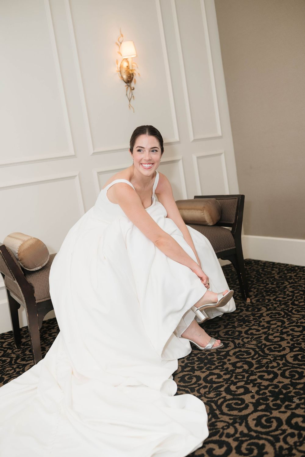 Smiling bride in white gown putting on silver heels while seated in an elegant hotel suite.