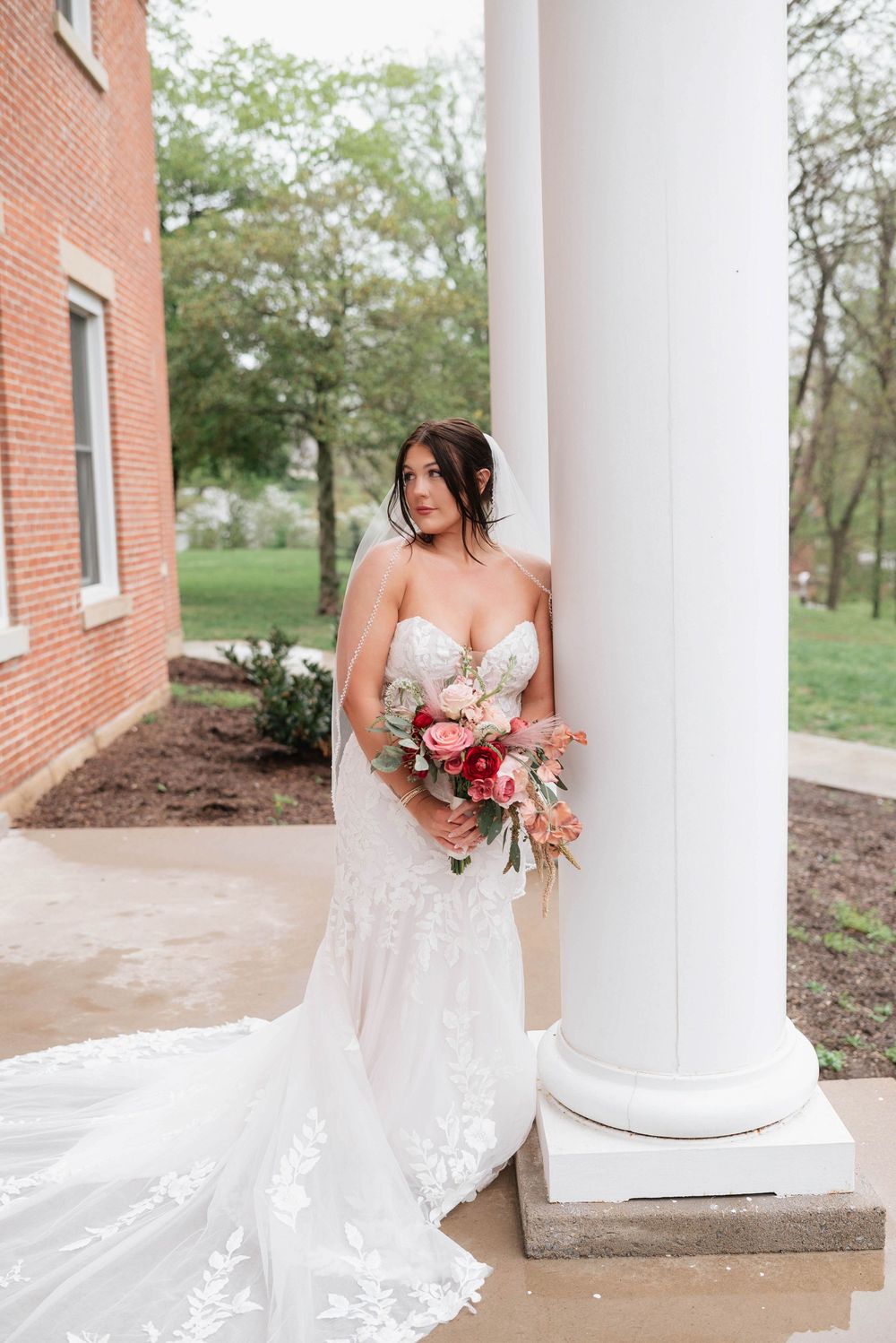 Bride in lace mermaid gown holding pink bouquet poses by white column of brick building outdoors.