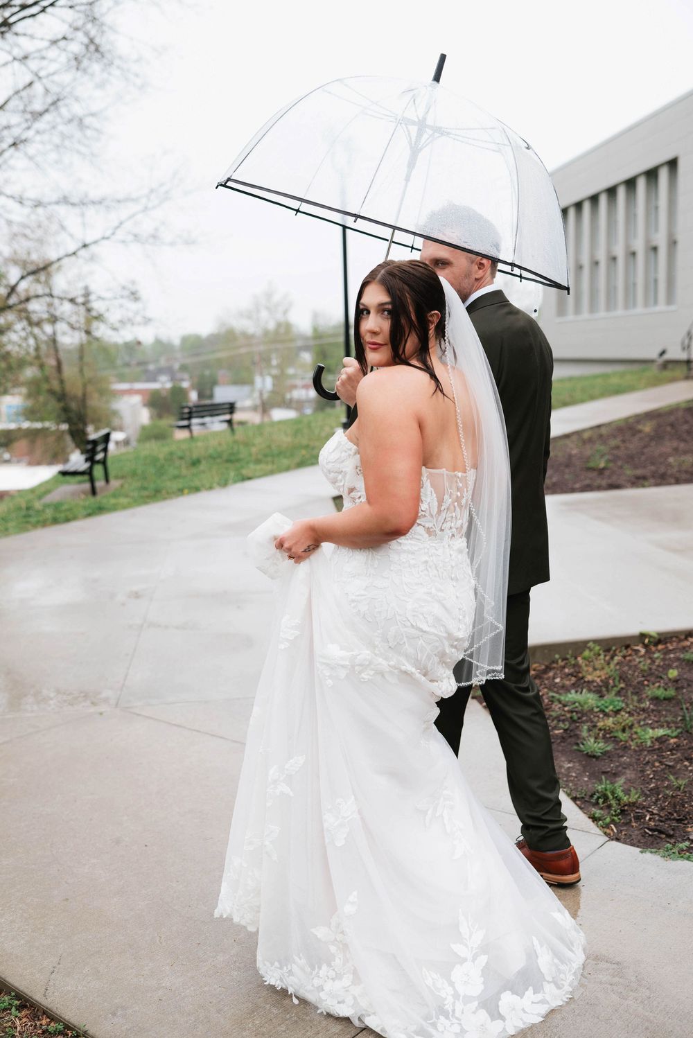 Bride in lace mermaid gown and groom sharing umbrella on rainy wedding day outdoor portrait.