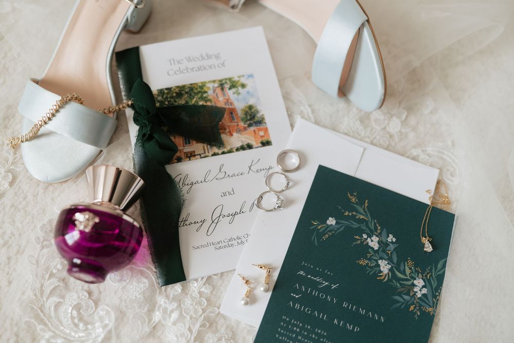 Elegant wedding flat lay with green invitation suite, gold rings, pearl earrings, and purple perfume bottle on lace fabric.