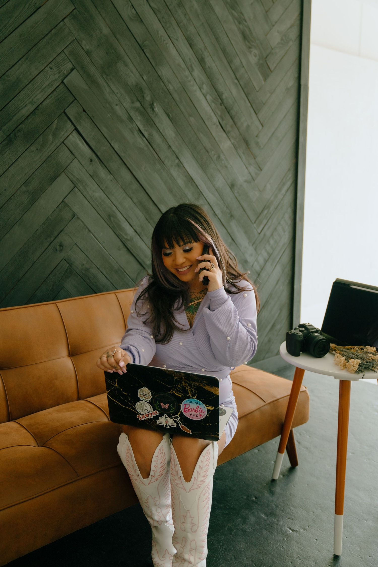 Professional with laptop and phone sitting on tan leather couch against dark herringbone wood wall.