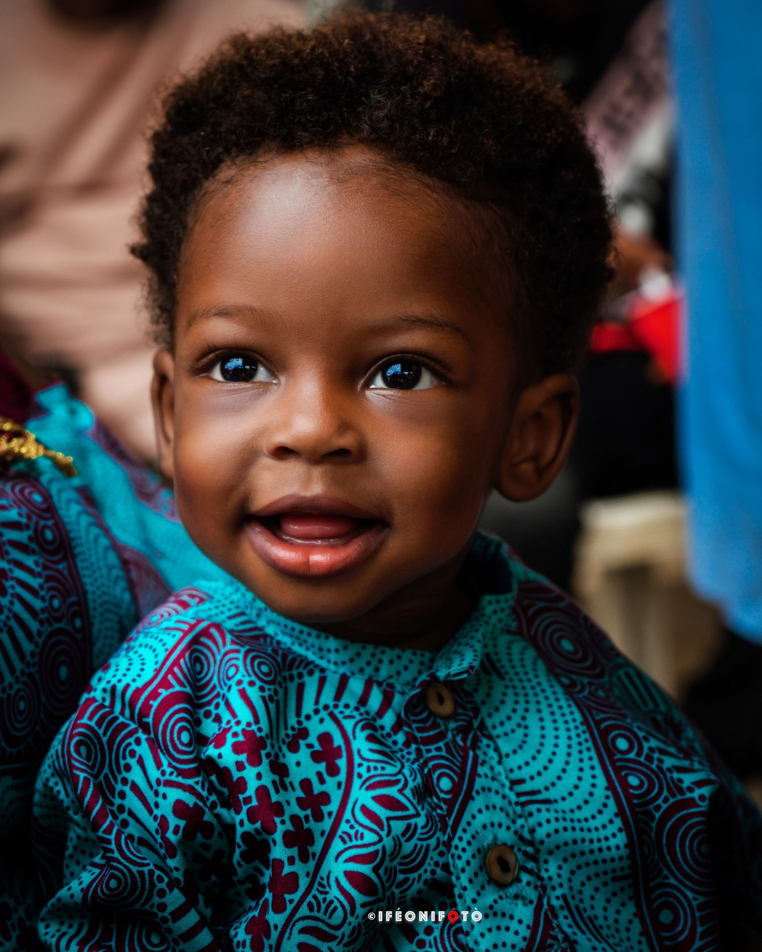 A charming baby boy with curly hair and bright eyes smiles joyfully in a portrait, wearing a vibrant blue and maroon African print outfit.
