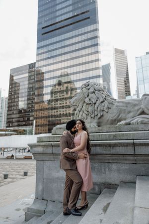 Couple embracing by stone lion statue in front of modern glass skyscraper building with reflective facade.