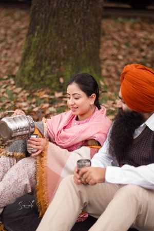 Two people enjoying a warm drink while sitting on fallen autumn leaves near a large tree.
