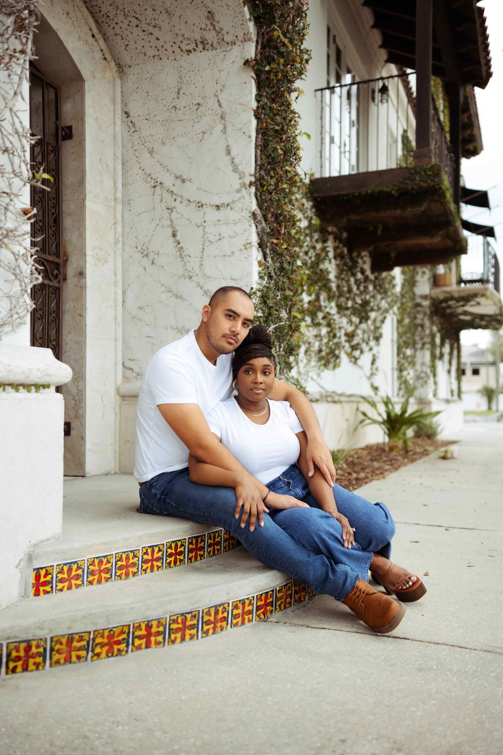 Couple sits together on decorative tiled steps in casual white outfits.