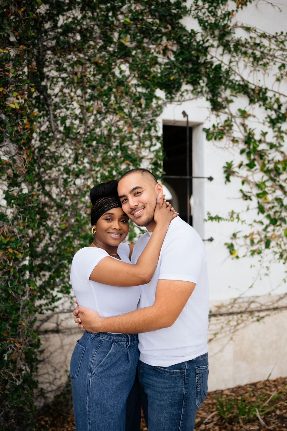 Couple in matching white t-shirts embracing outdoors near white building with climbing vines.