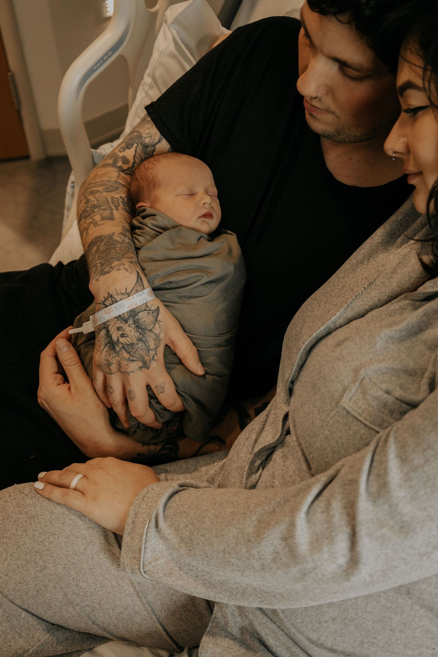 New parents with tattoos tenderly hold their swaddled newborn baby together in a hospital room.