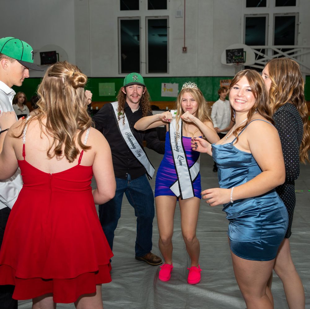 A group of partygoers in formal dresses dance together at an indoor event with a sash-wearing attendee in the center.