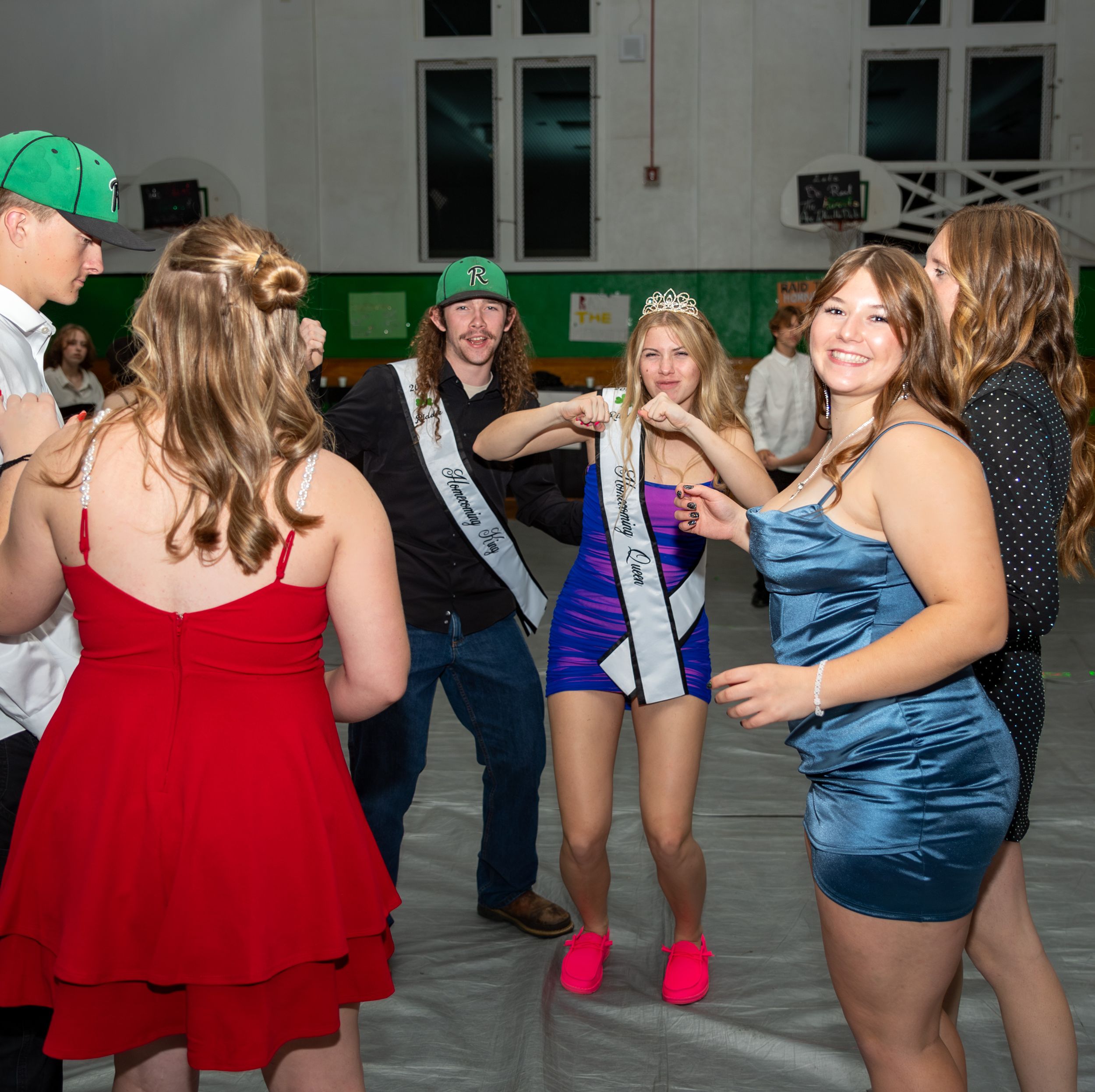 A group of partygoers in formal dresses dance together at an indoor event with a sash-wearing attendee in the center.