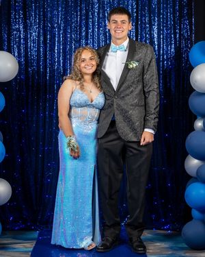 A couple poses at prom in front of a sparkly blue backdrop with balloon decorations, wearing blue and black formal attire.