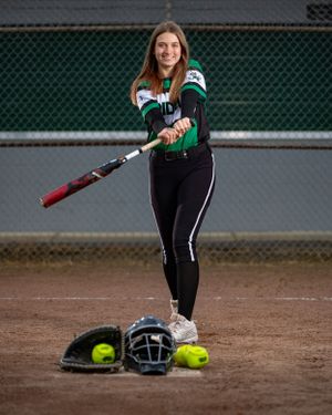 A softball player in uniform poses with a bat and equipment on a dirt field.