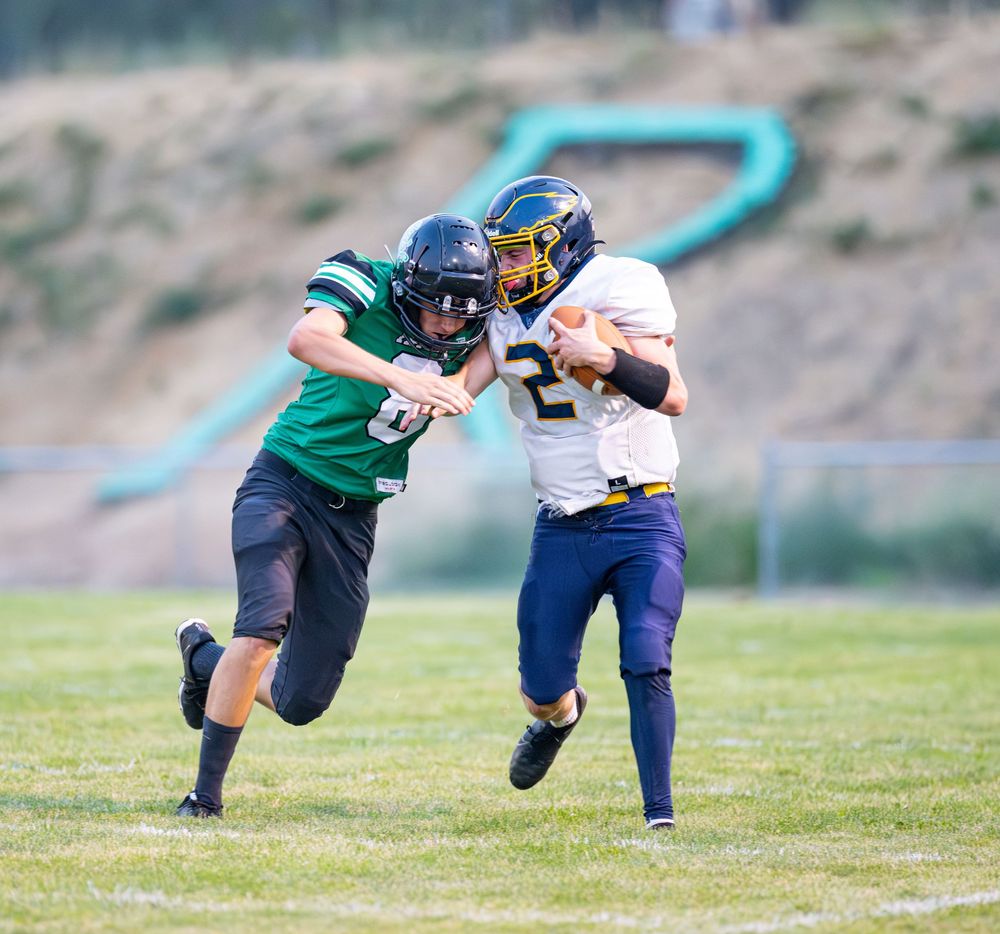 Two football players collide during a high school game on a grassy field with one in green and one in white uniforms.