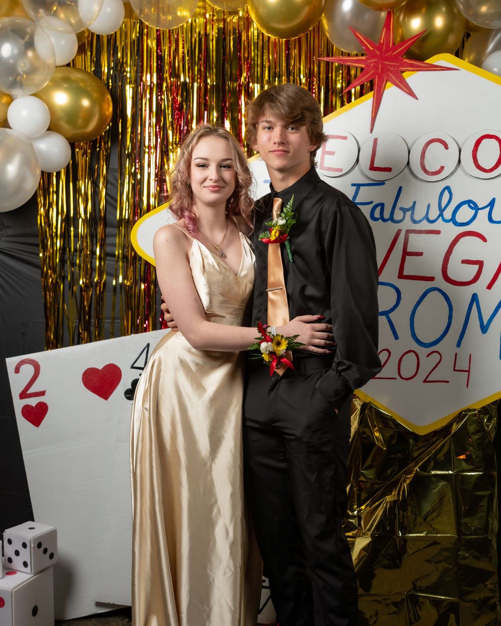 Couple in formal prom attire pose against gold streamers and casino-themed party decorations.