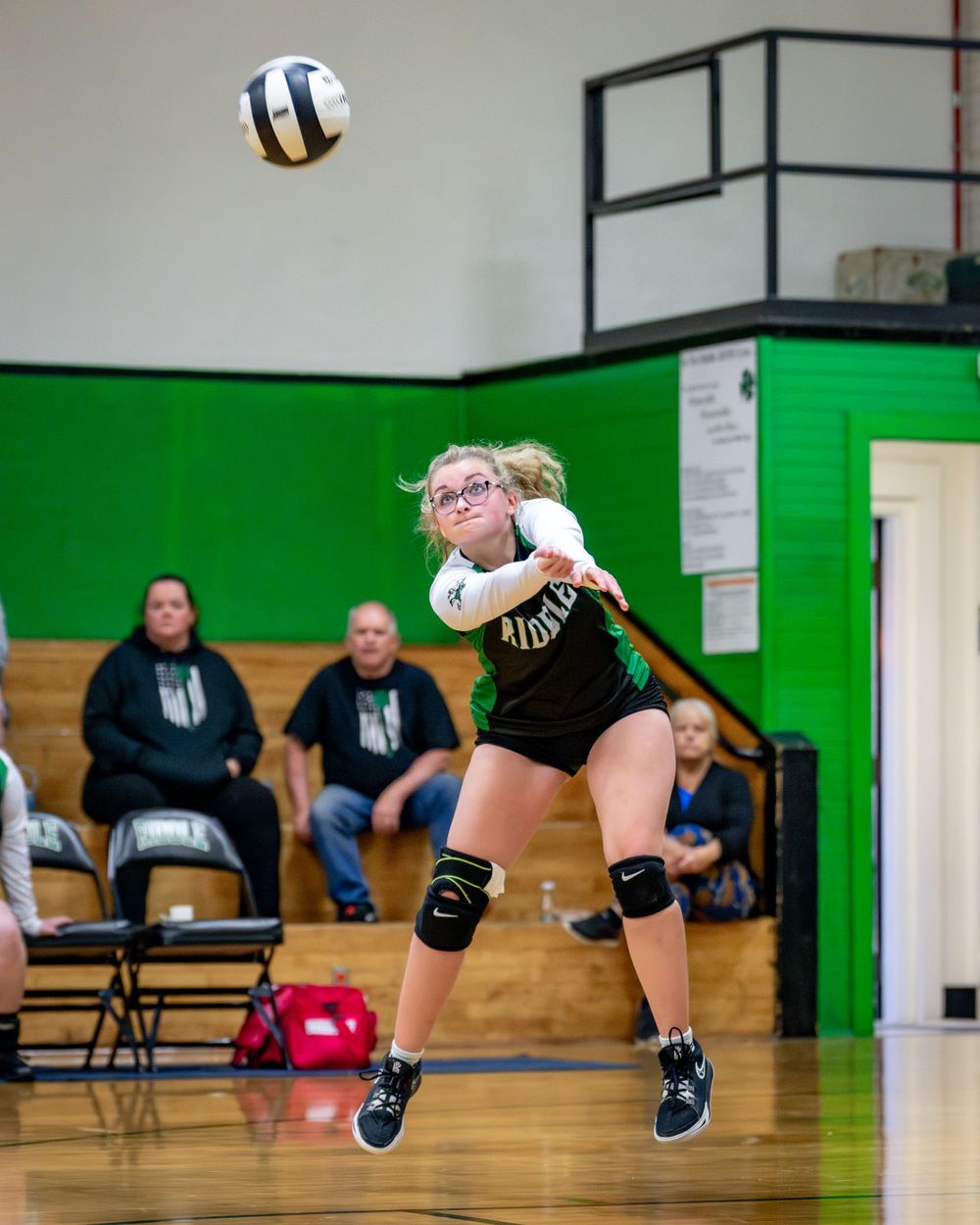 A volleyball player in a black uniform jumps to return a serve during a match in a green-walled gymnasium.