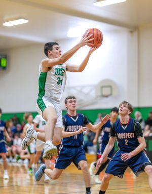 High school basketball player in white jersey jumps for layup while two defenders in navy uniforms watch below.