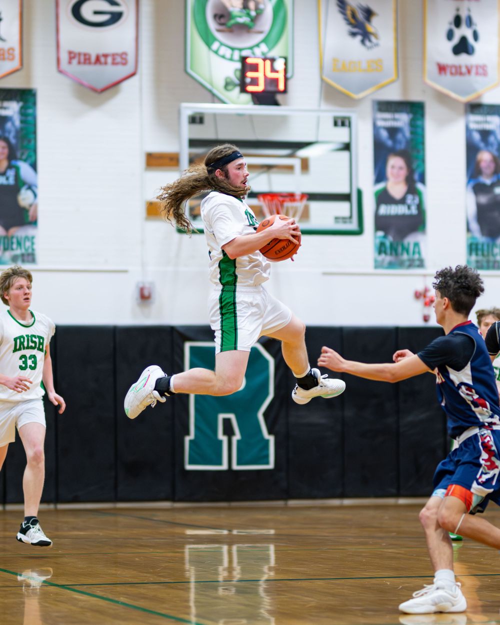 Basketball player leaps high in the air while handling the ball during an indoor game with school banners in background.
