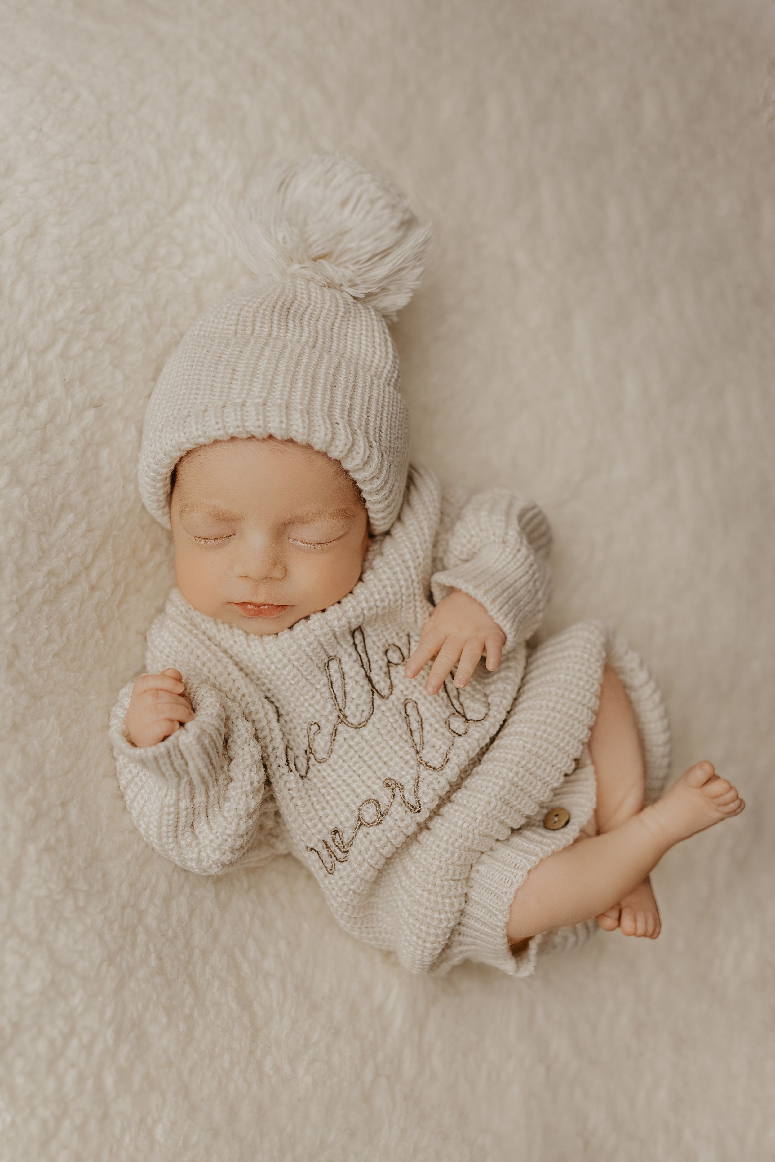 Sleeping newborn wearing a cozy white knit outfit on a cream-colored textured background.