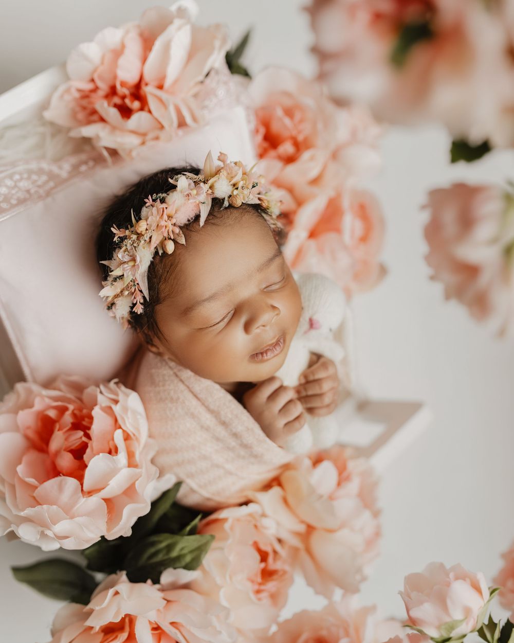 Newborn baby sleeping peacefully surrounded by soft peach roses and delicate floral arrangements.