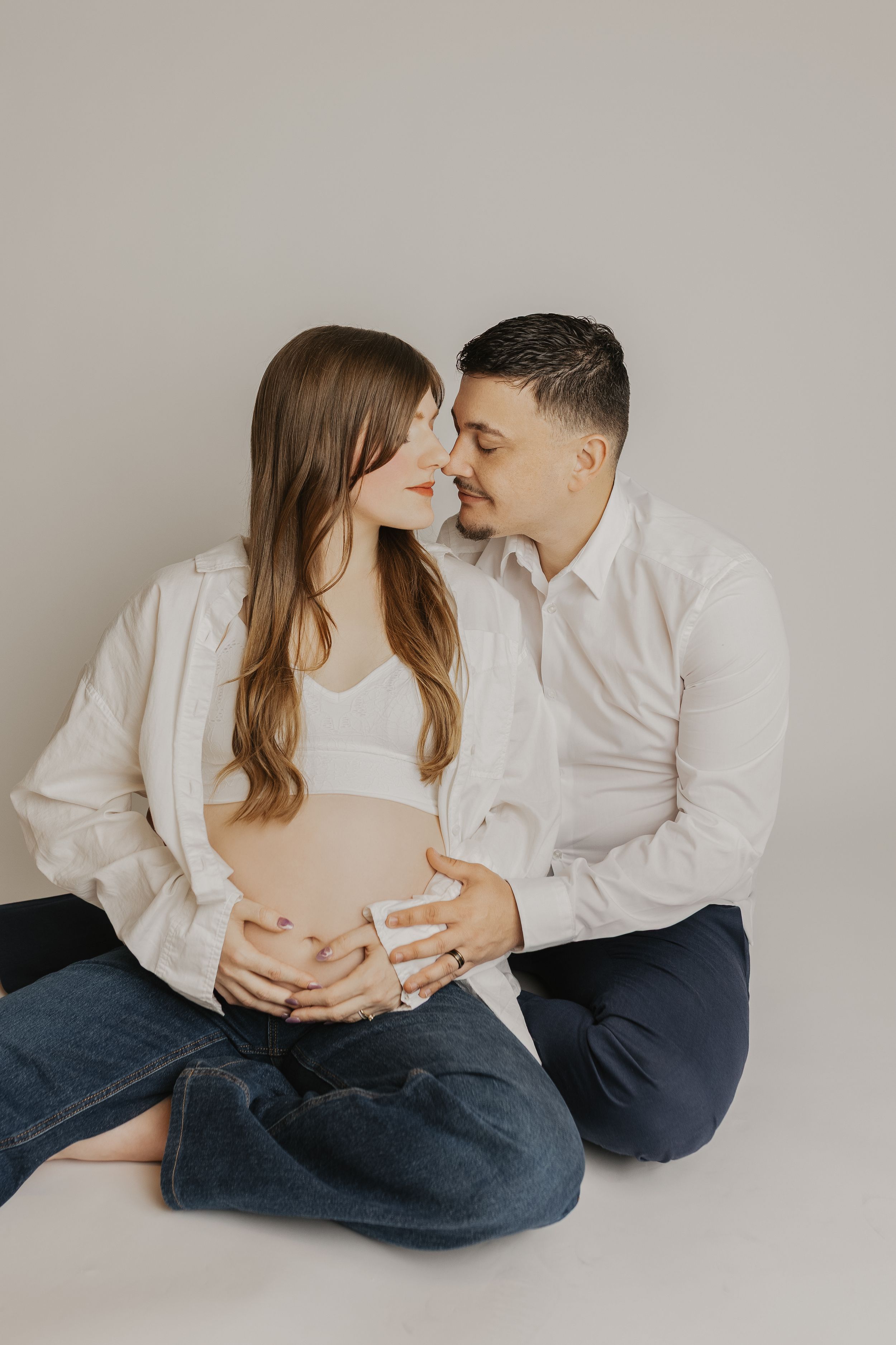Romantic maternity photo series showing couple in white shirts and jeans sharing intimate moments on light background.