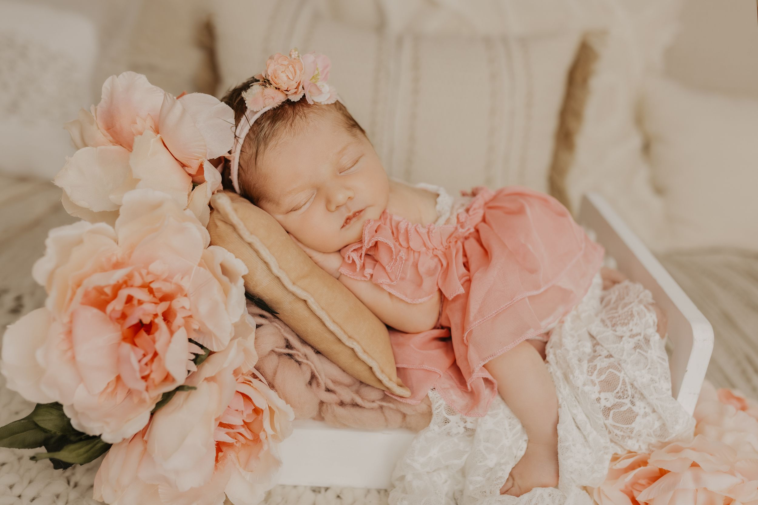 Newborn baby girl sleeping peacefully in pink ruffle outfit surrounded by soft peach peonies in a white wooden crib.