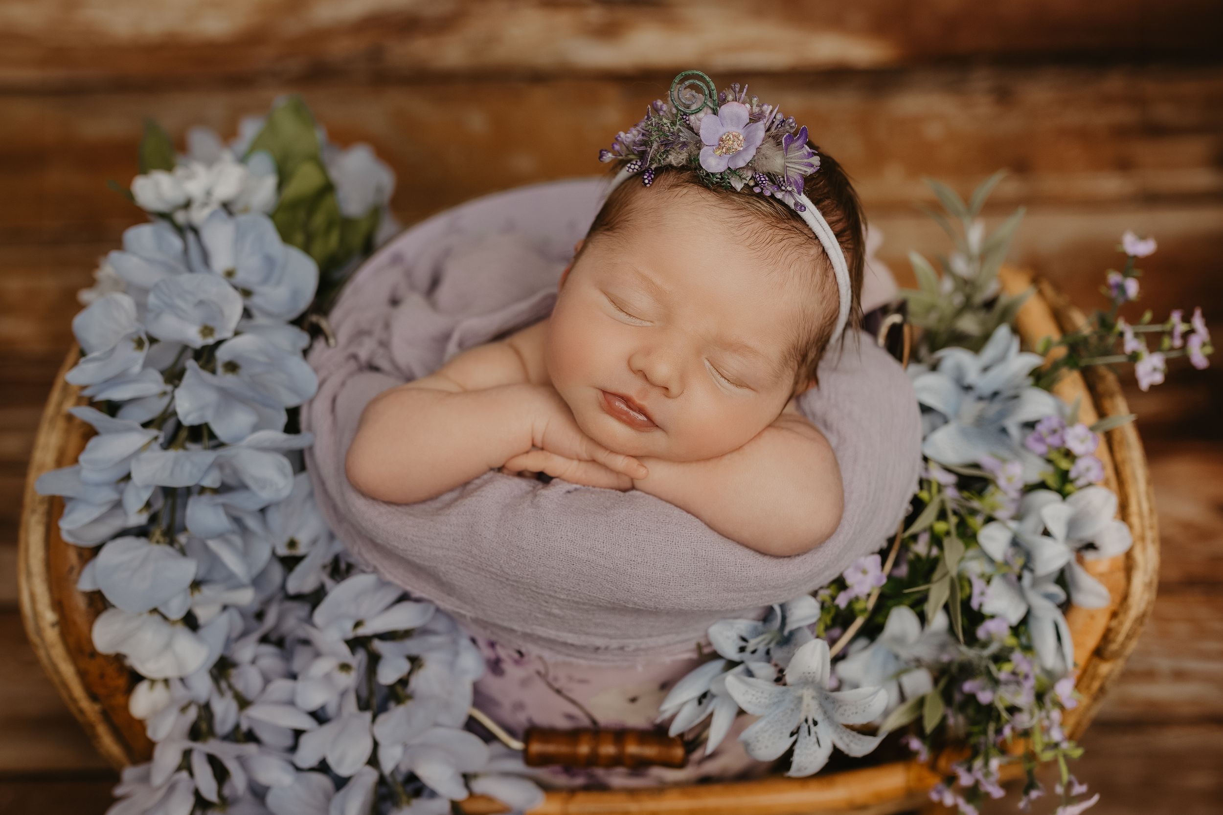 Newborn baby girl sleeping peacefully in a basket surrounded by soft blue flowers, wearing a delicate floral headband.