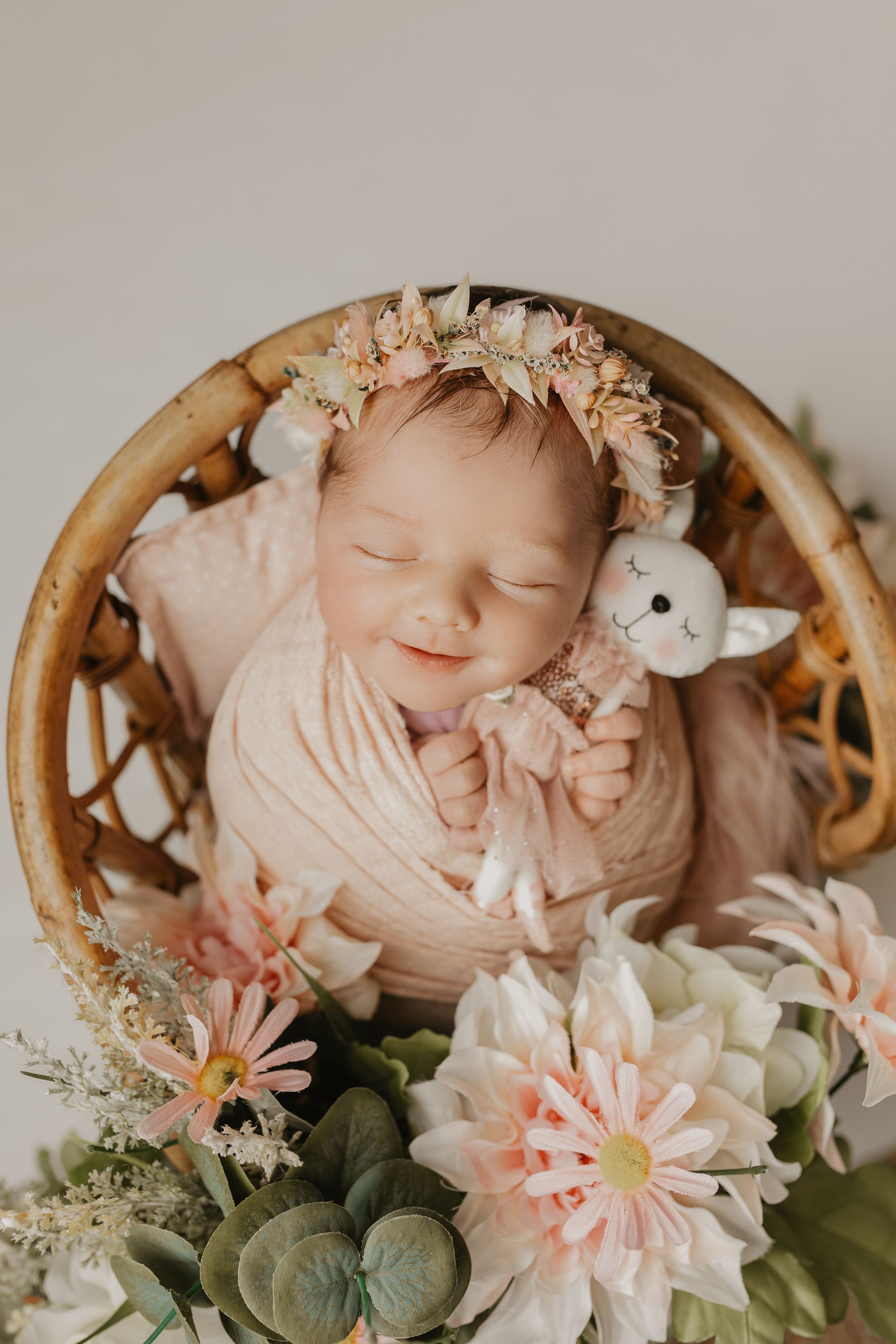 Sleeping newborn baby wrapped in pink, wearing floral headband, holding bunny toy in wicker basket with flowers.