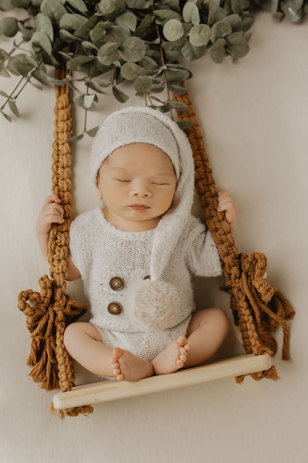 Sleeping newborn baby in white knit romper and bonnet on macrame swing with eucalyptus leaves.