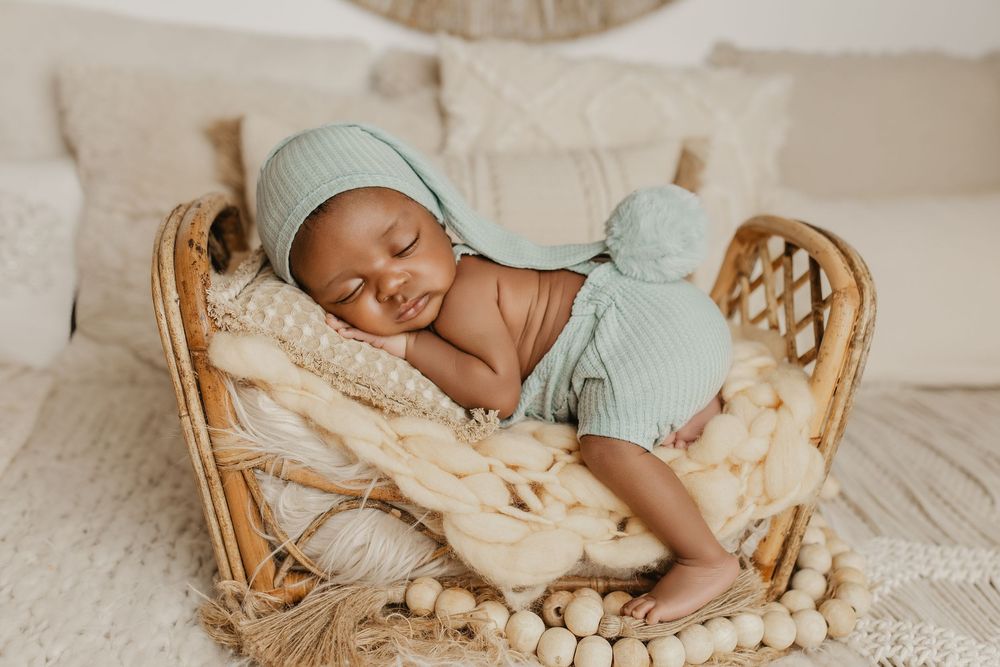 Newborn baby sleeping peacefully in a wicker basket, wearing a sage green knit outfit and hat.