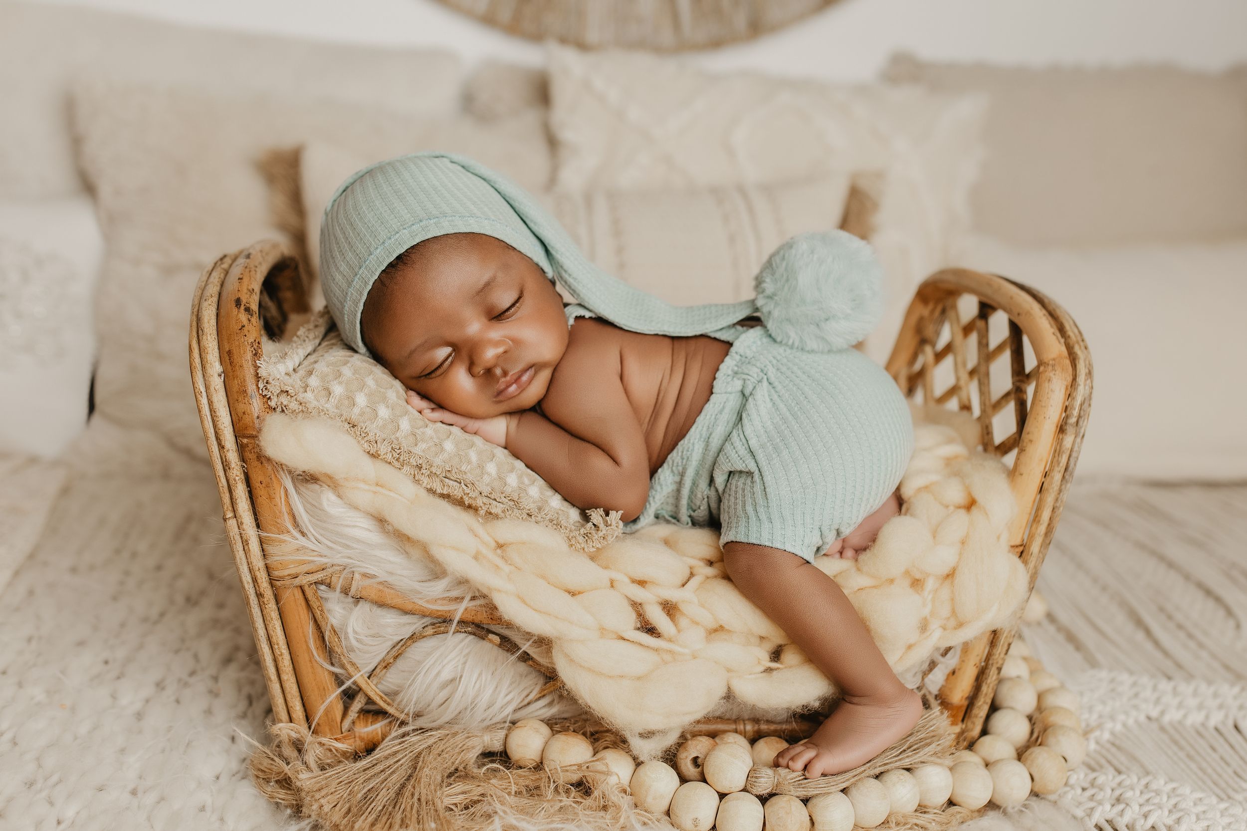 Newborn baby sleeping peacefully in a wicker basket, wearing a sage green knit outfit and hat.